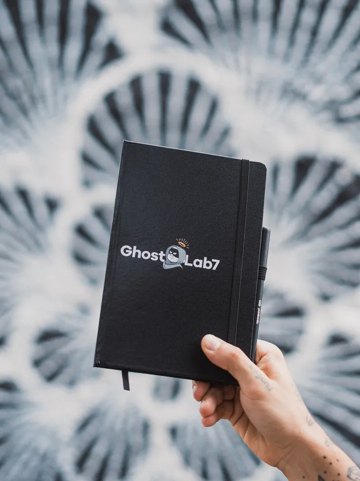 Una mano sostiene el cuaderno negro de Ghost Lab7 frente a un mural en blanco y negro