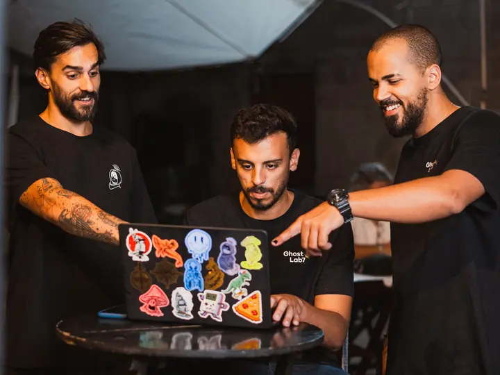 Tres personas del equipo de Ghost Lab7 riendo alrededor de una laptop llena de stickers